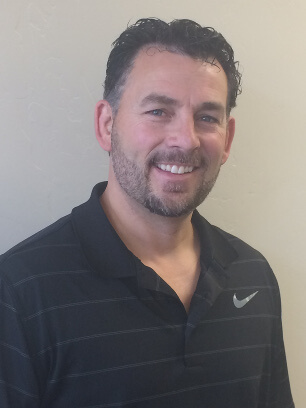 A man with short dark curly hair and a trimmed beard smiles at the camera. He is wearing a black collared shirt with a subtle stripe pattern and a gray Nike logo on the chest, standing against a plain light-colored background.
