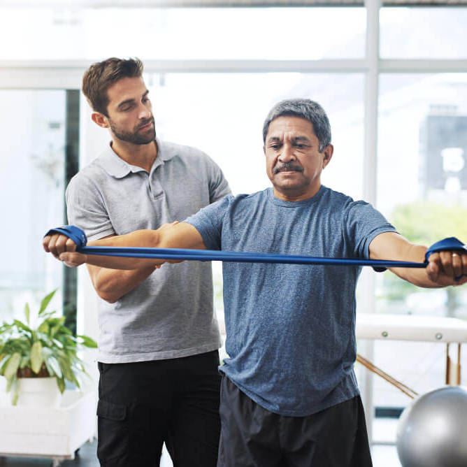 Senior man performing physical therapy exercises with resistance band under supervision.